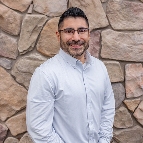 Man with glasses and dark hair smiling against a stone wall