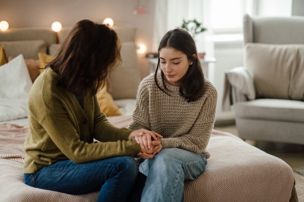 Two women sit on a bed holding hands, one offering comfort as the other looks down with a thoughtful expression.
