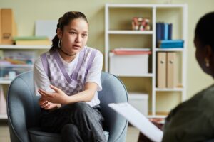 Teenager sitting in a chair talks with a counselor, appearing thoughtful during a conversation in an office setting.