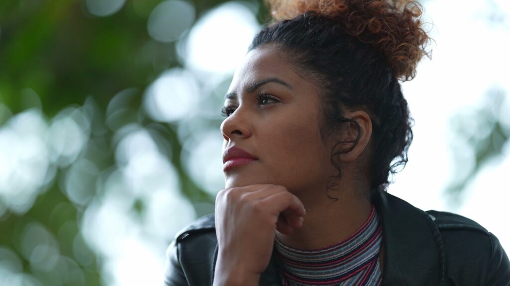 Woman sitting outdoors with her chin resting on her hand, looking thoughtful and pensive. She has curly hair tied up and wears a striped top and black jacket, with blurred greenery in the background.