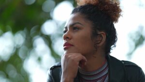 Woman sitting outdoors with her chin resting on her hand, looking thoughtful and pensive. She has curly hair tied up and wears a striped top and black jacket, with blurred greenery in the background.